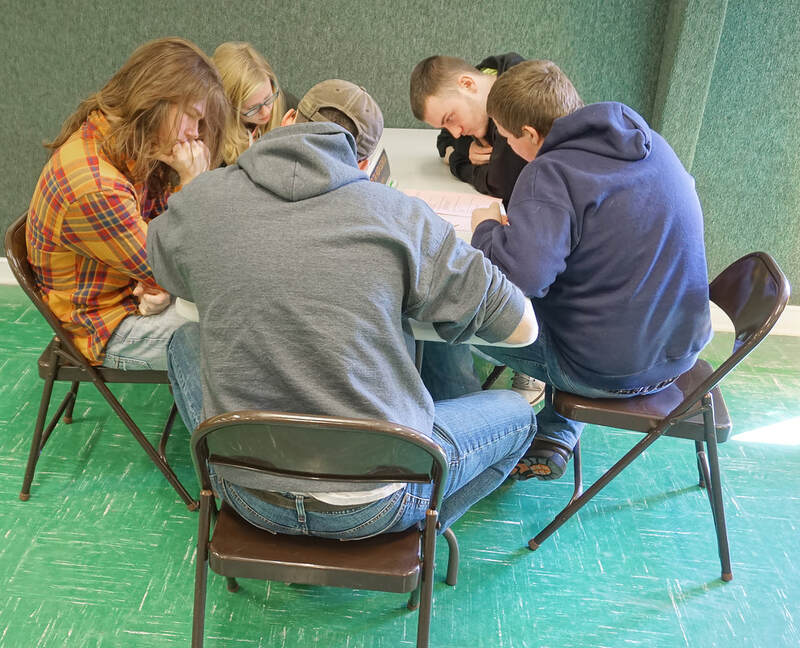 Five students are seated around a table focusing intently on a paper test. 